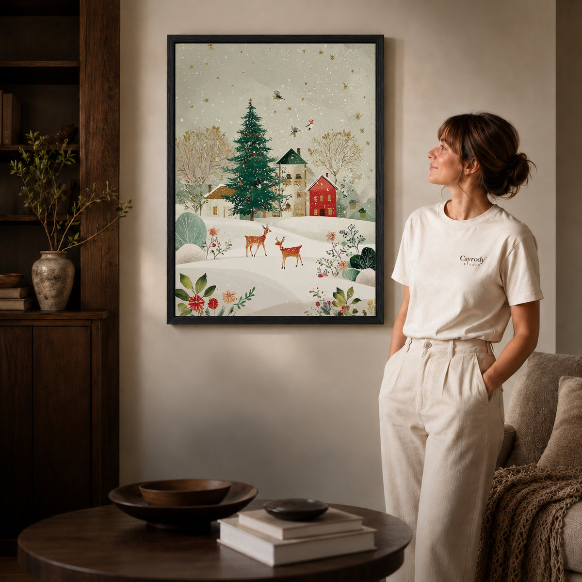 snowy village deer canvas art in a black frame beside a woman and round wooden table.