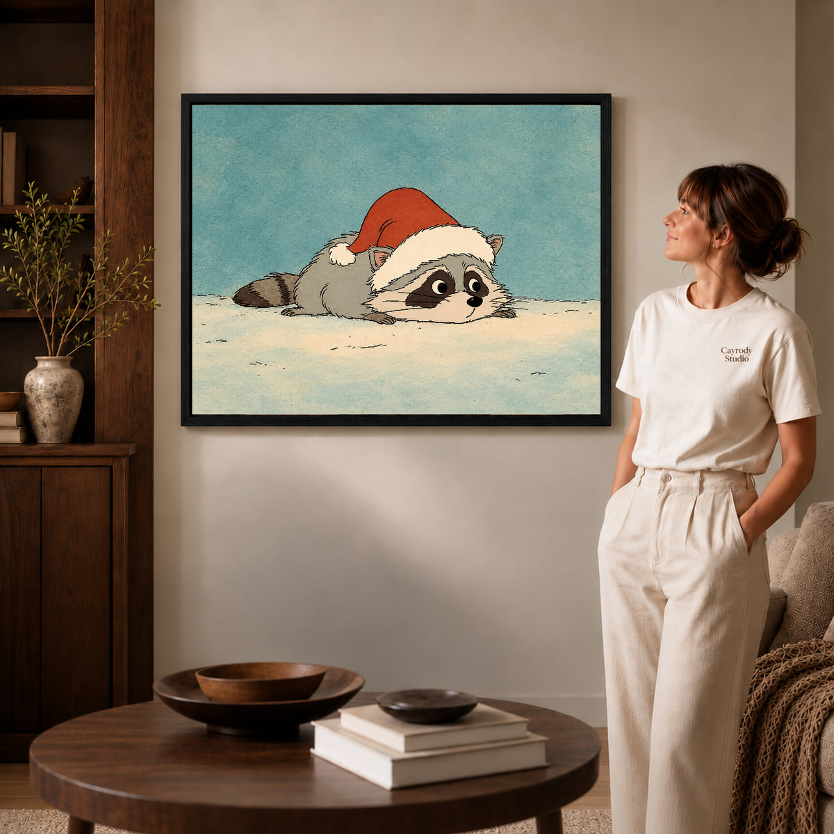 sleepy holiday raccoon canvas art in a black frame beside a woman and round wooden table.