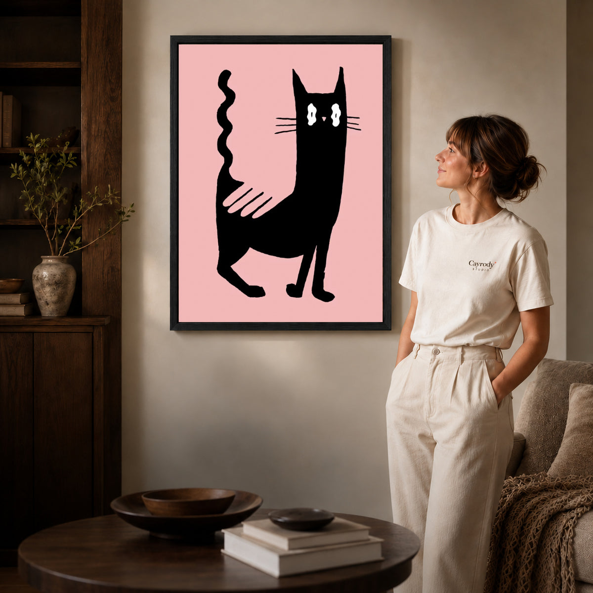 wavy tail cat canvas art in a black frame beside a woman and round wooden table.