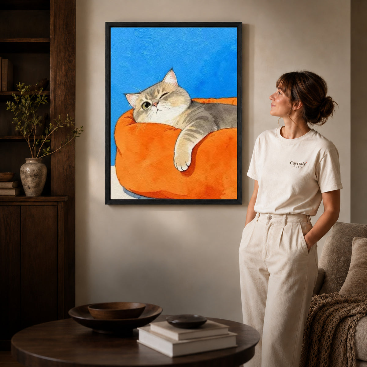 winking armchair cat canvas art in a black frame beside a woman and round wooden table.