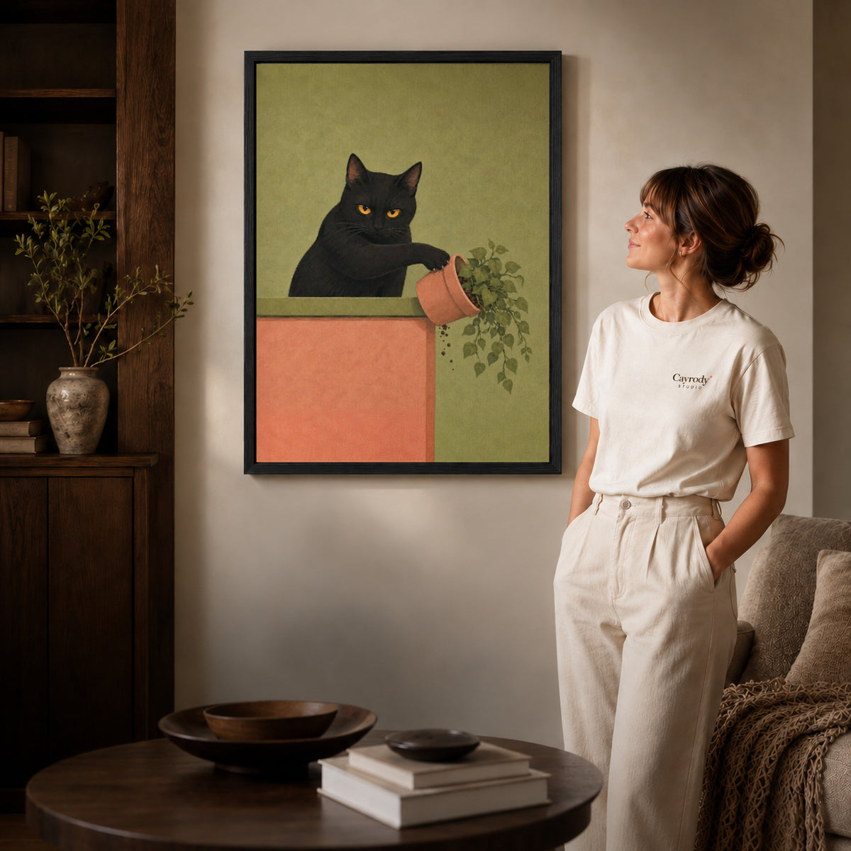 mischievous plant cat canvas art in a black frame beside a woman and round wooden table.