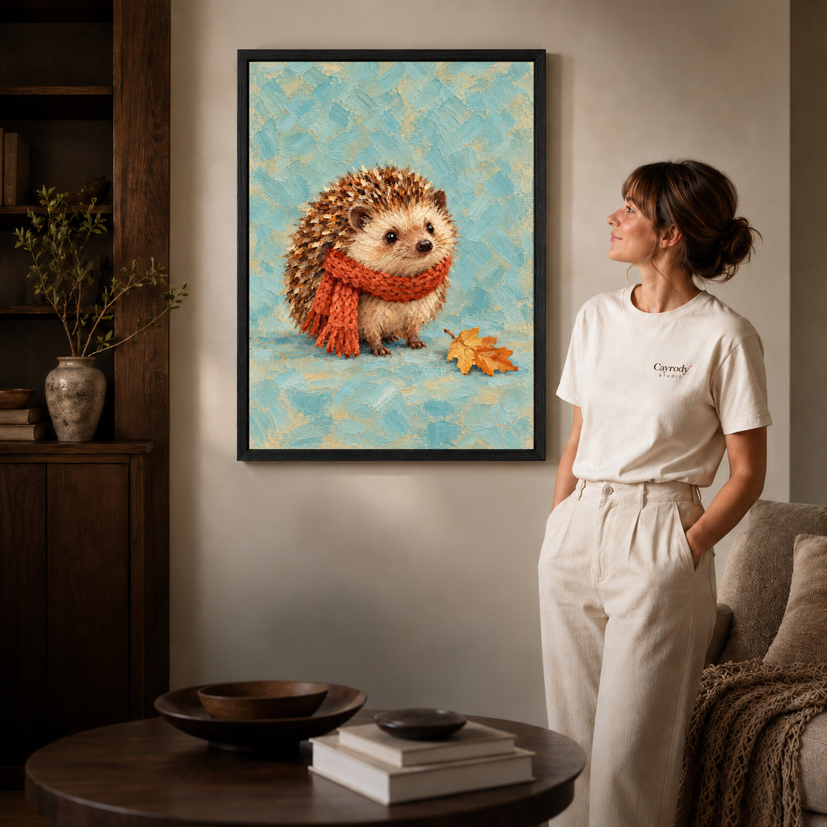 autumn scarf hedgehog canvas art in a black frame beside a woman and round wooden table.