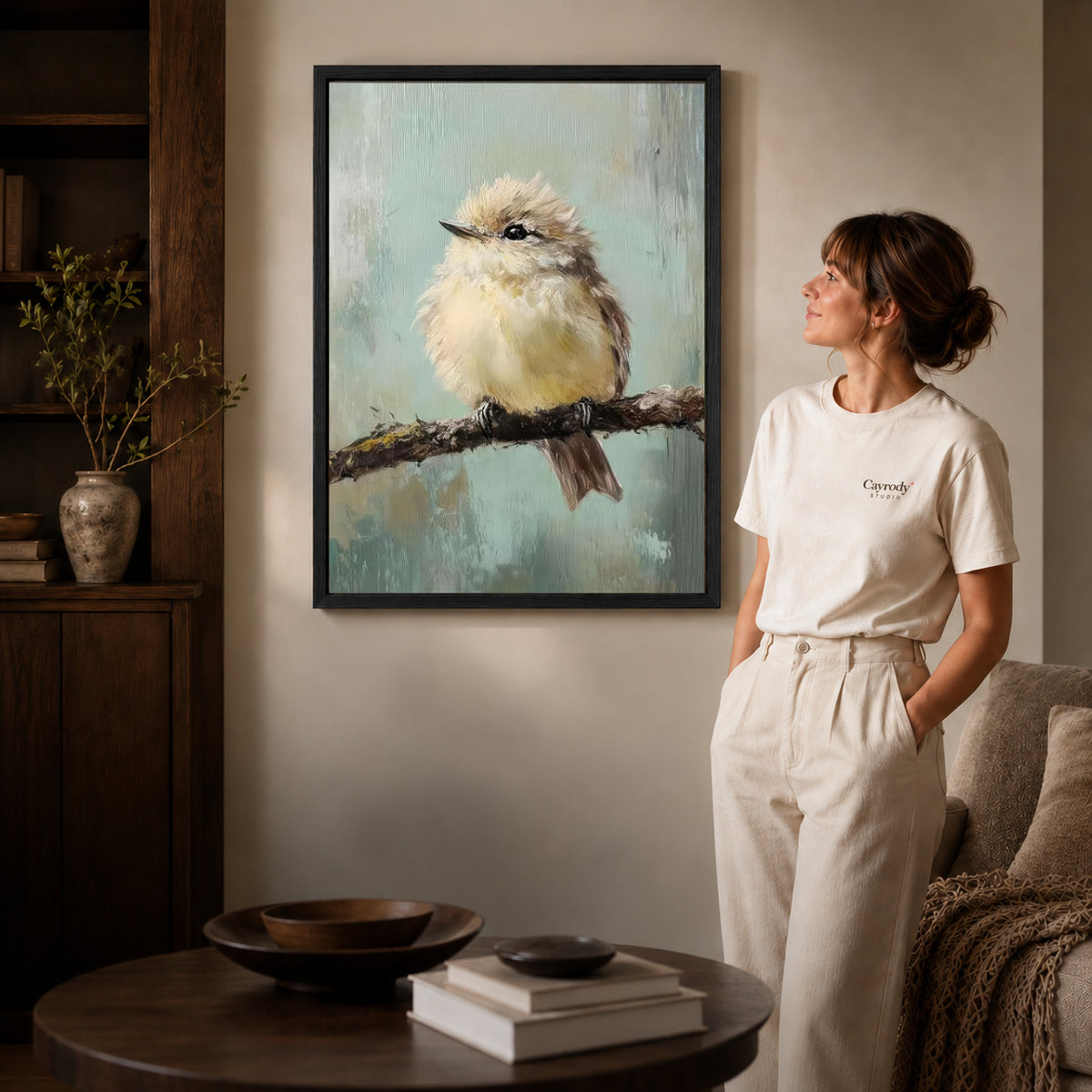little branch companion canvas art in a black frame beside a woman and round wooden table.