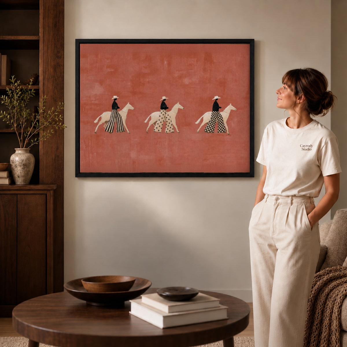 patterned rider parade canvas art in a black frame beside a woman and round wooden table.
