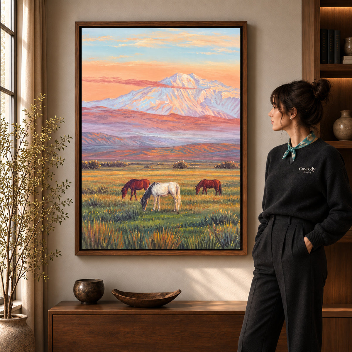 pastel mountain horses canvas art in a walnut frame beside a standing woman and wooden console.