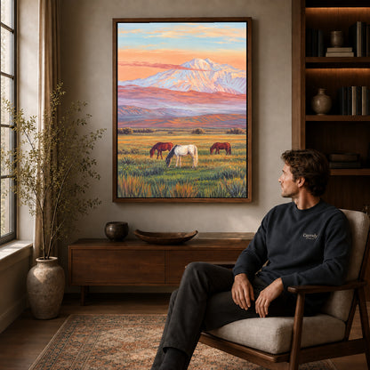 pastel mountain horses canvas art in a walnut frame above a console beside a seated man.