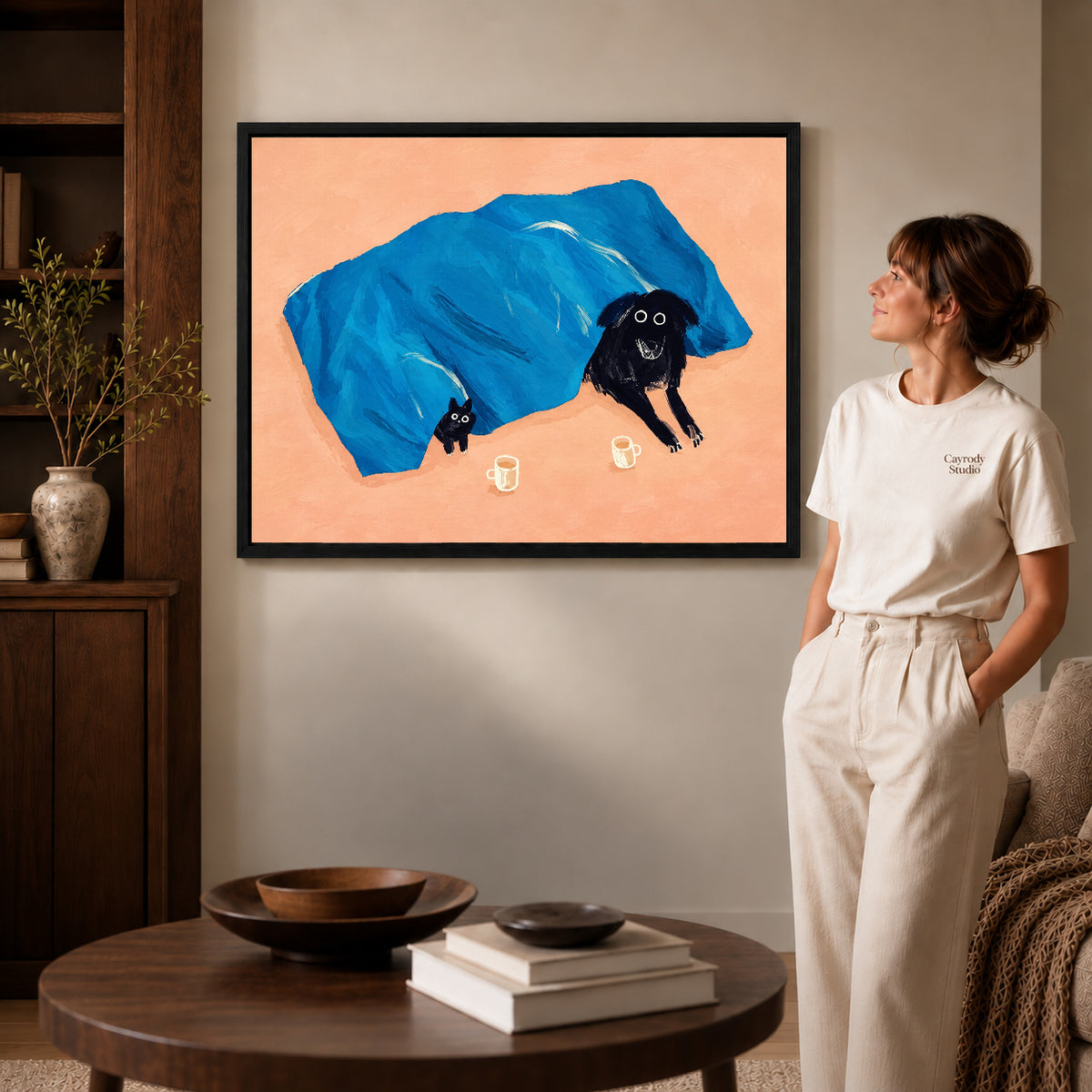 cozy blanket companions canvas art in a black frame beside a woman and round wooden table.
