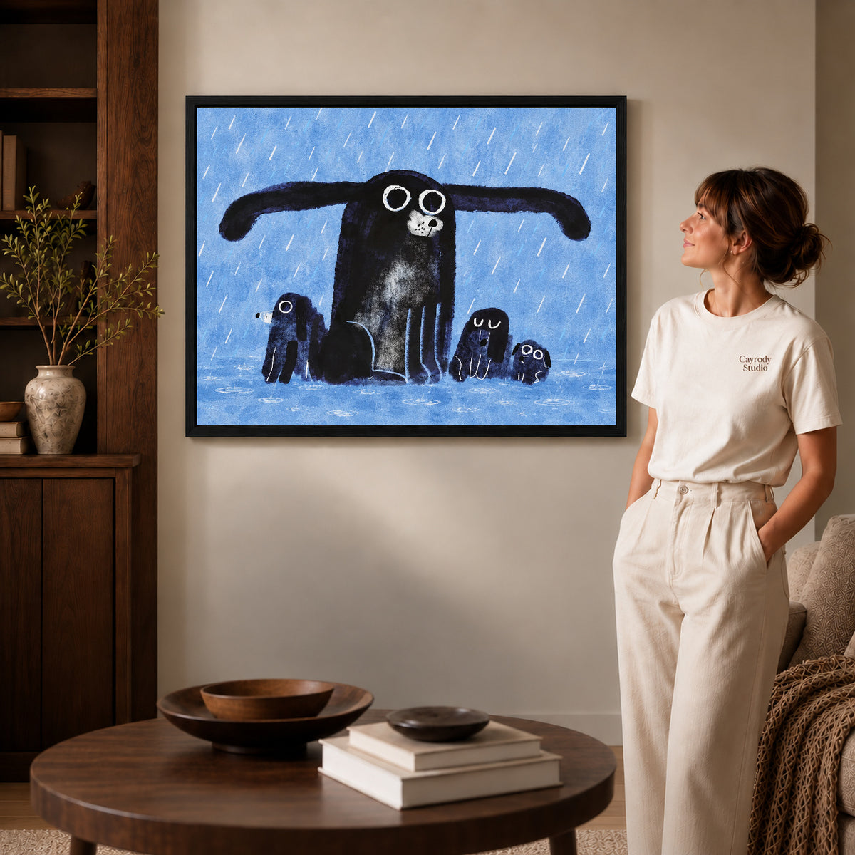 sheltering dog family canvas art in a black frame beside a woman and round wooden table.