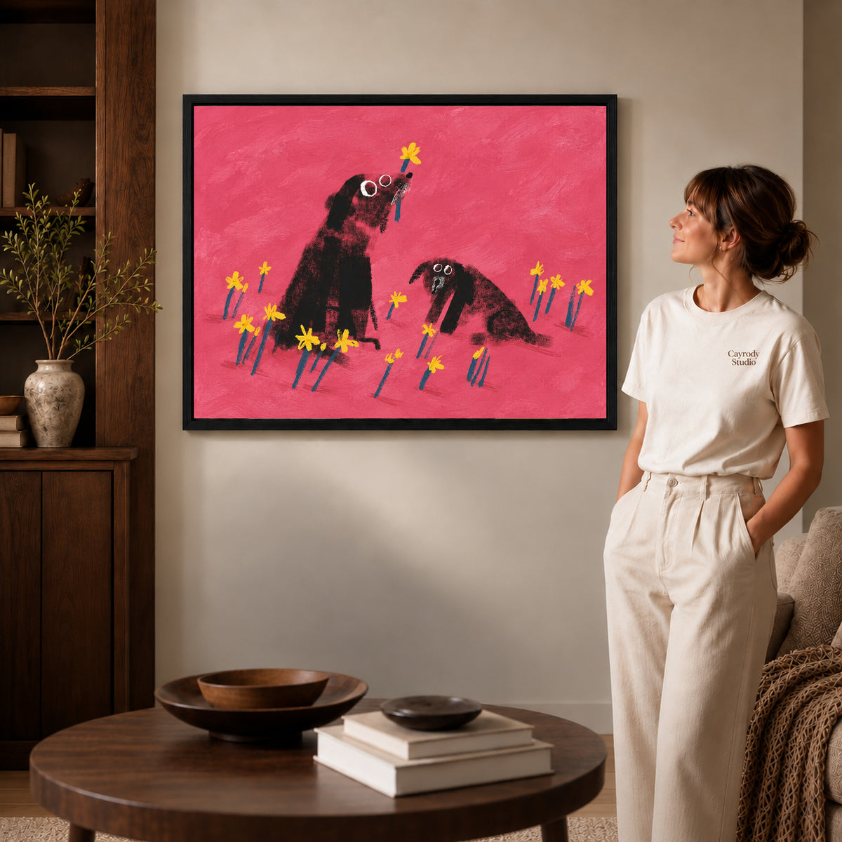 dogs among daffodils canvas art in a black frame beside a woman and round wooden table.