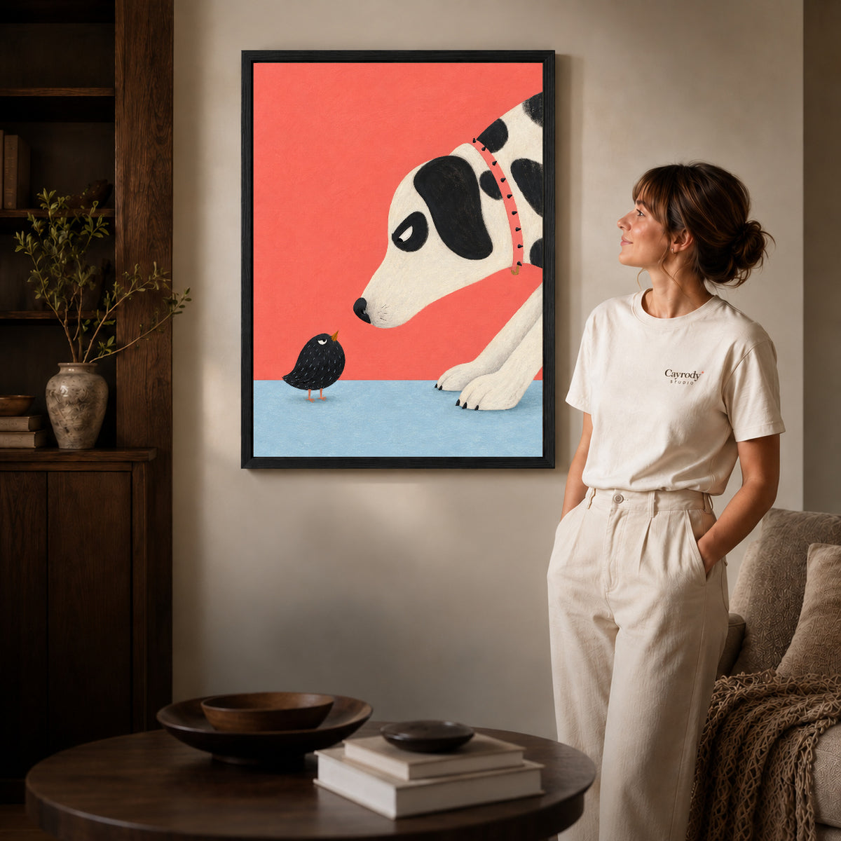 dog meets tiny bird canvas art in a black frame beside a woman and round wooden table.