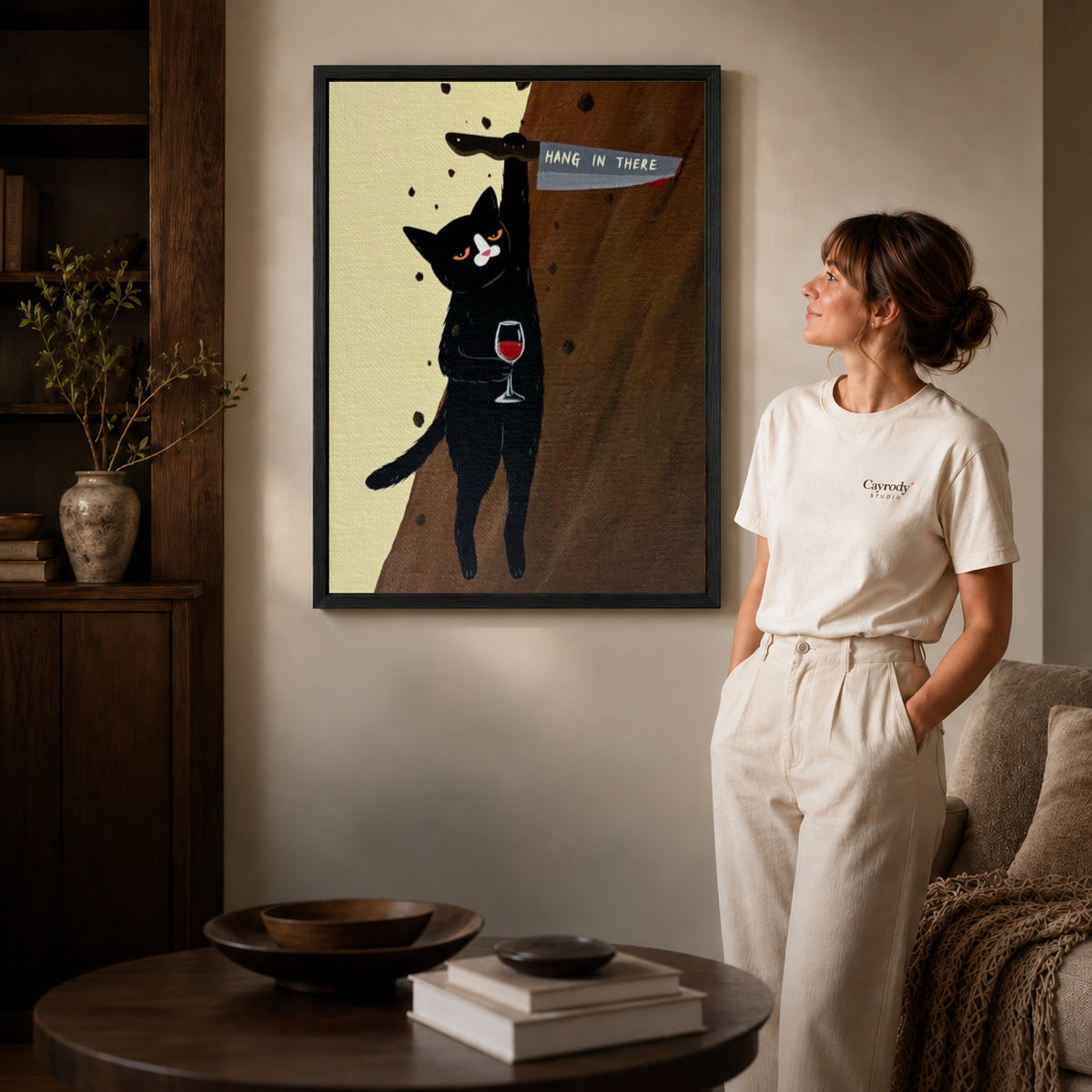 hang in there cat canvas art in a black frame beside a woman and round wooden table.