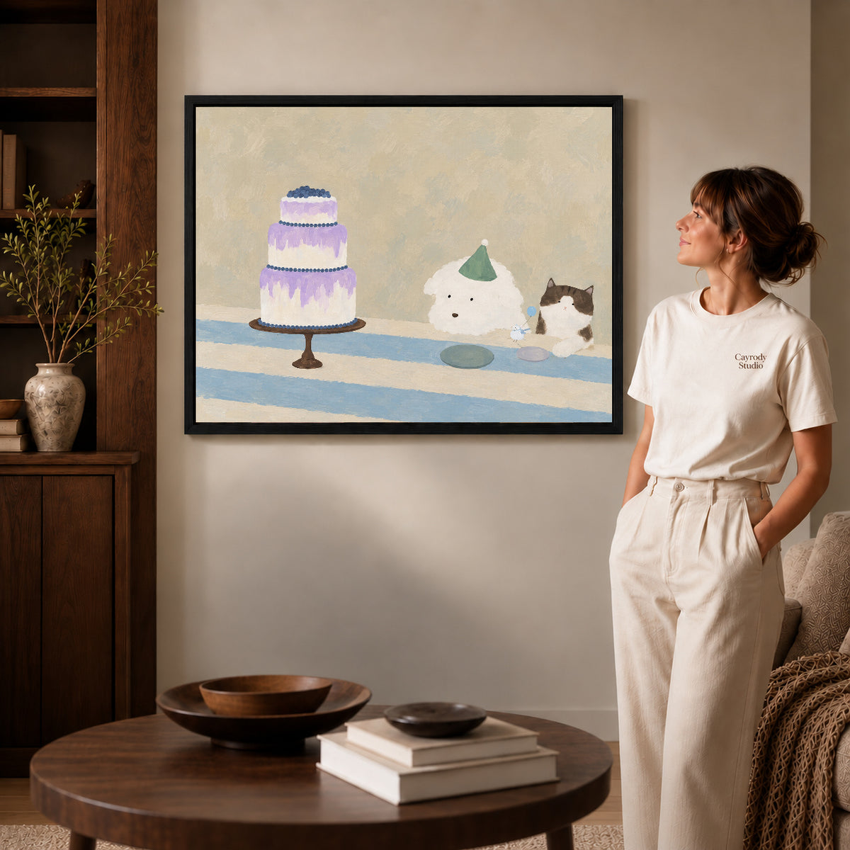 cake party companions canvas art in a black frame beside a woman and round wooden table.