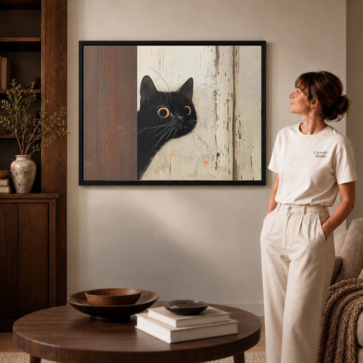 Black framed peeking black cat canvas art displayed beside a woman in a warm neutral living room.