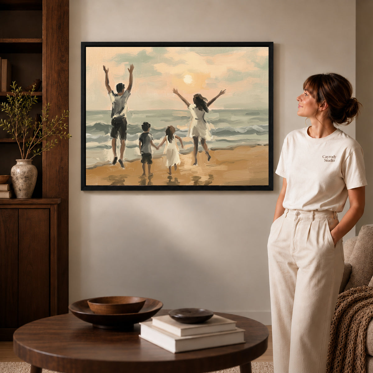 Black framed family beach sunset canvas art displayed beside a woman in a warm neutral living room.