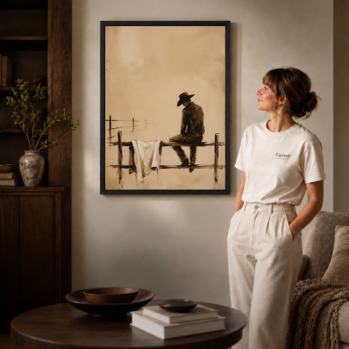 Black framed sepia cowboy canvas art displayed beside a woman in a warm neutral living room.