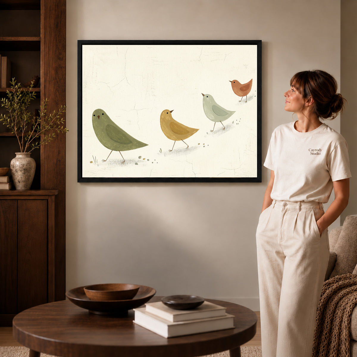 Black framed muted folk birds canvas art displayed beside a woman in a warm neutral living room.