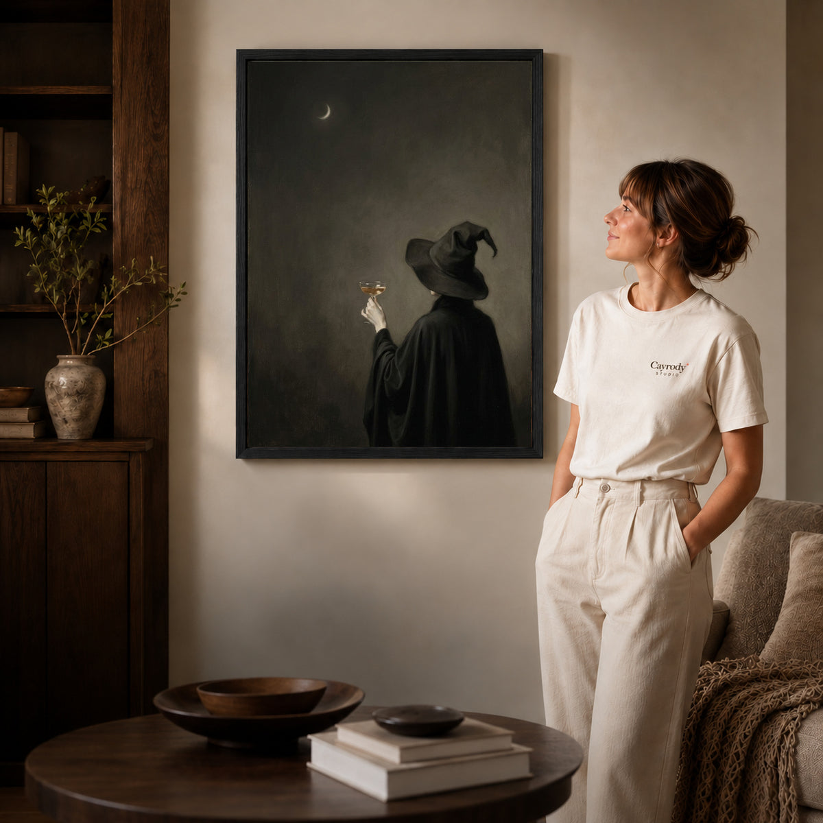 Black framed moody witch toast canvas art displayed beside a woman in a warm neutral living room.