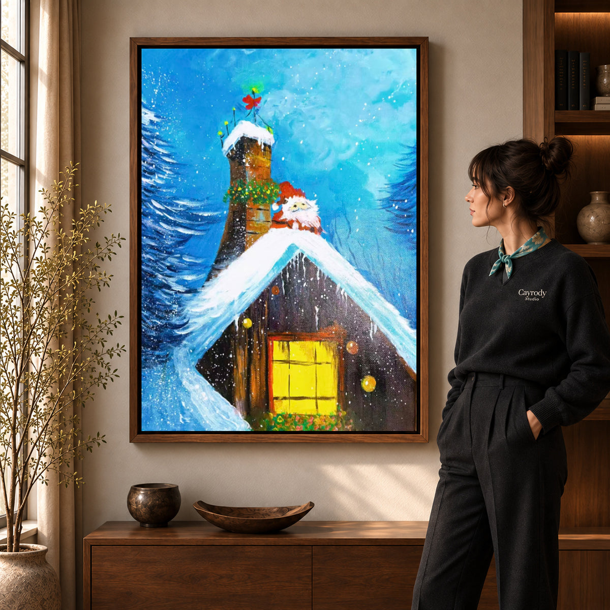 Snowy cottage canvas art in a walnut frame beside a standing woman in a warm living room.