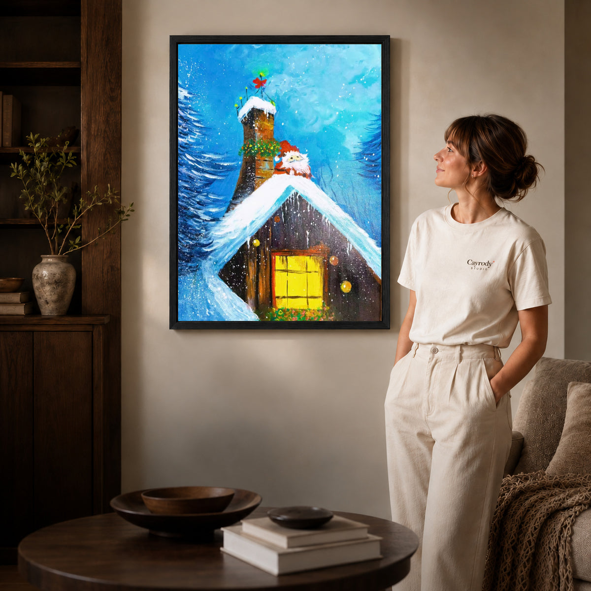 Black framed snowy cottage canvas art displayed beside a woman in a warm neutral living room.