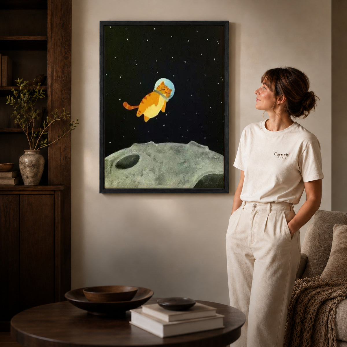 Black framed space kitten canvas art displayed beside a woman in a warm neutral living room.