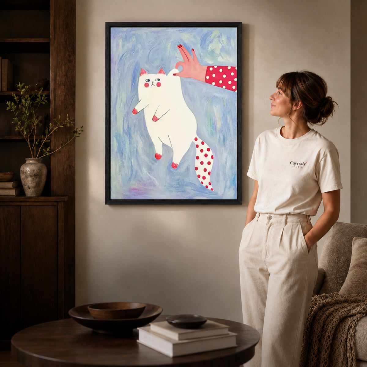 Black framed playful white cat canvas art displayed beside a woman in a warm neutral living room.