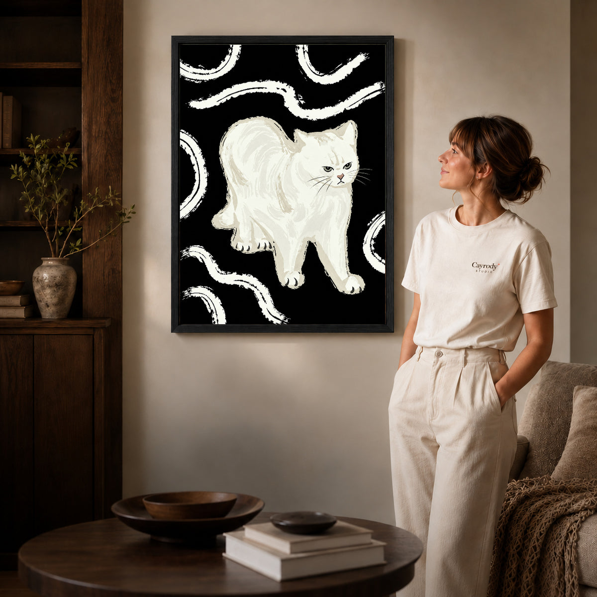 Black framed grumpy white cat artwork beside a woman in a warm neutral living room.