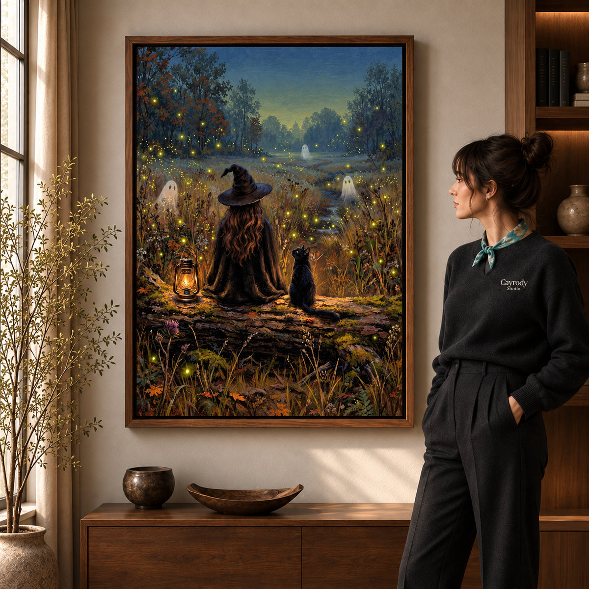 Witch cat fireflies canvas art in a walnut frame beside a standing woman in a warm living room.