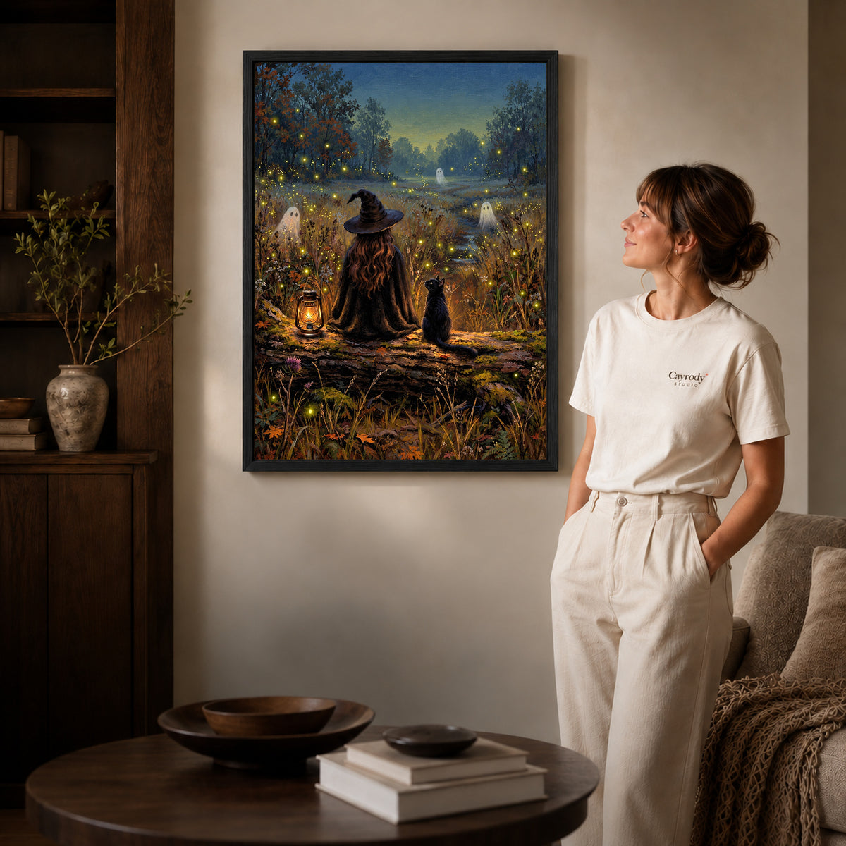 Black framed witch cat fireflies canvas art displayed beside a woman in a warm neutral living room.
