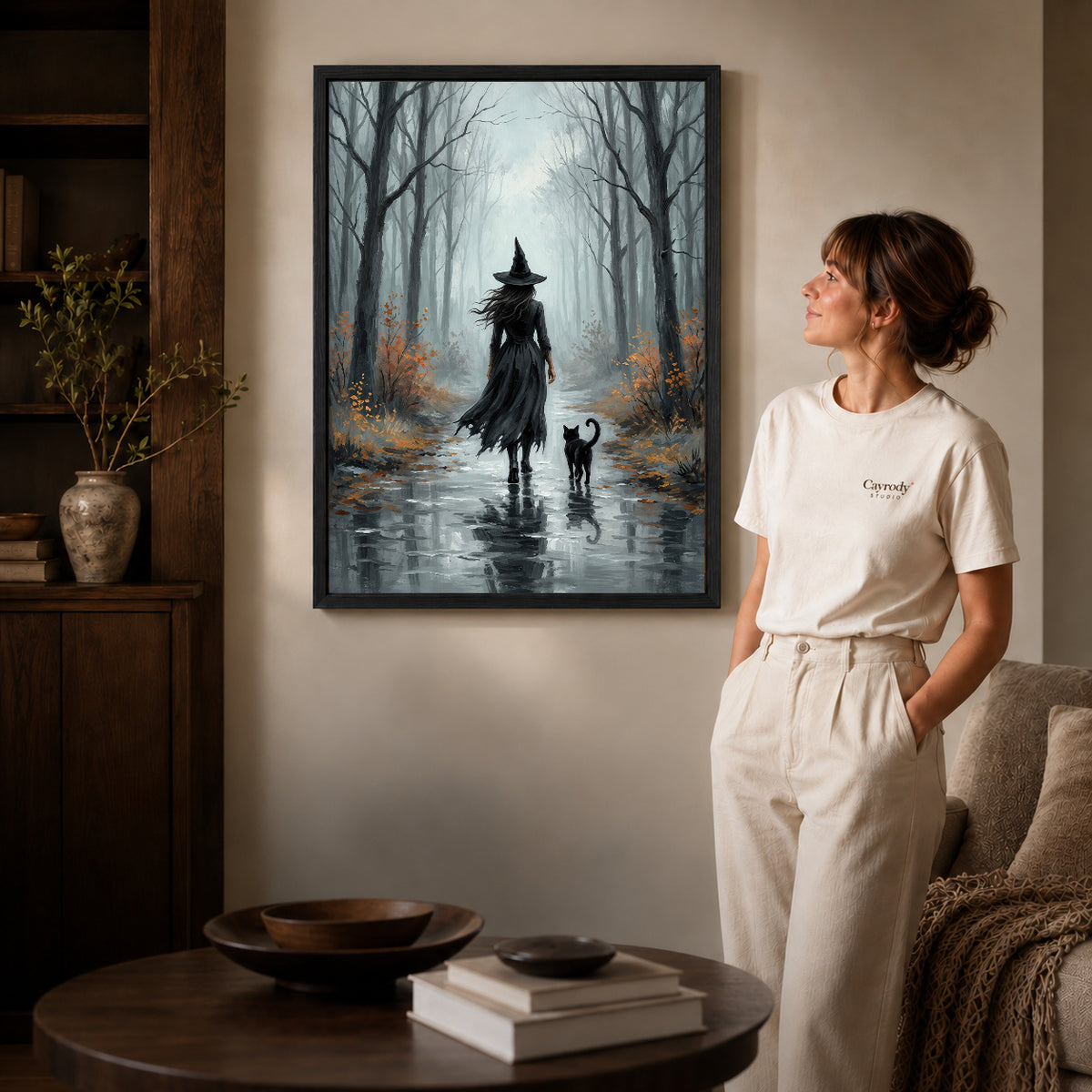 Black framed misty witch walk artwork beside a woman in a warm neutral living room.