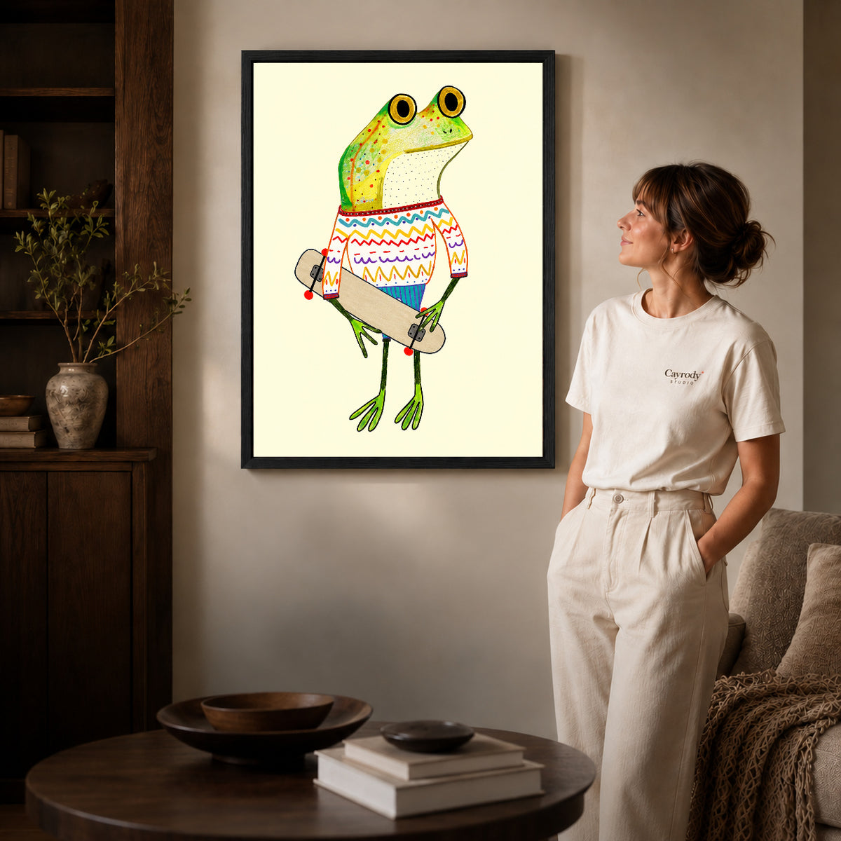 Black framed skateboard sweater frog artwork beside a woman in a warm neutral living room.