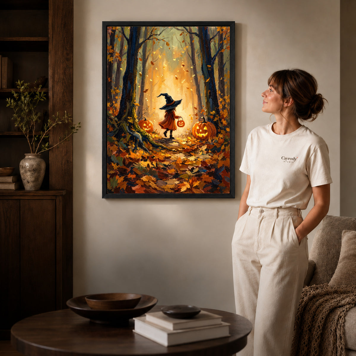 Black framed autumn witch path canvas art displayed beside a woman in a warm neutral living room.