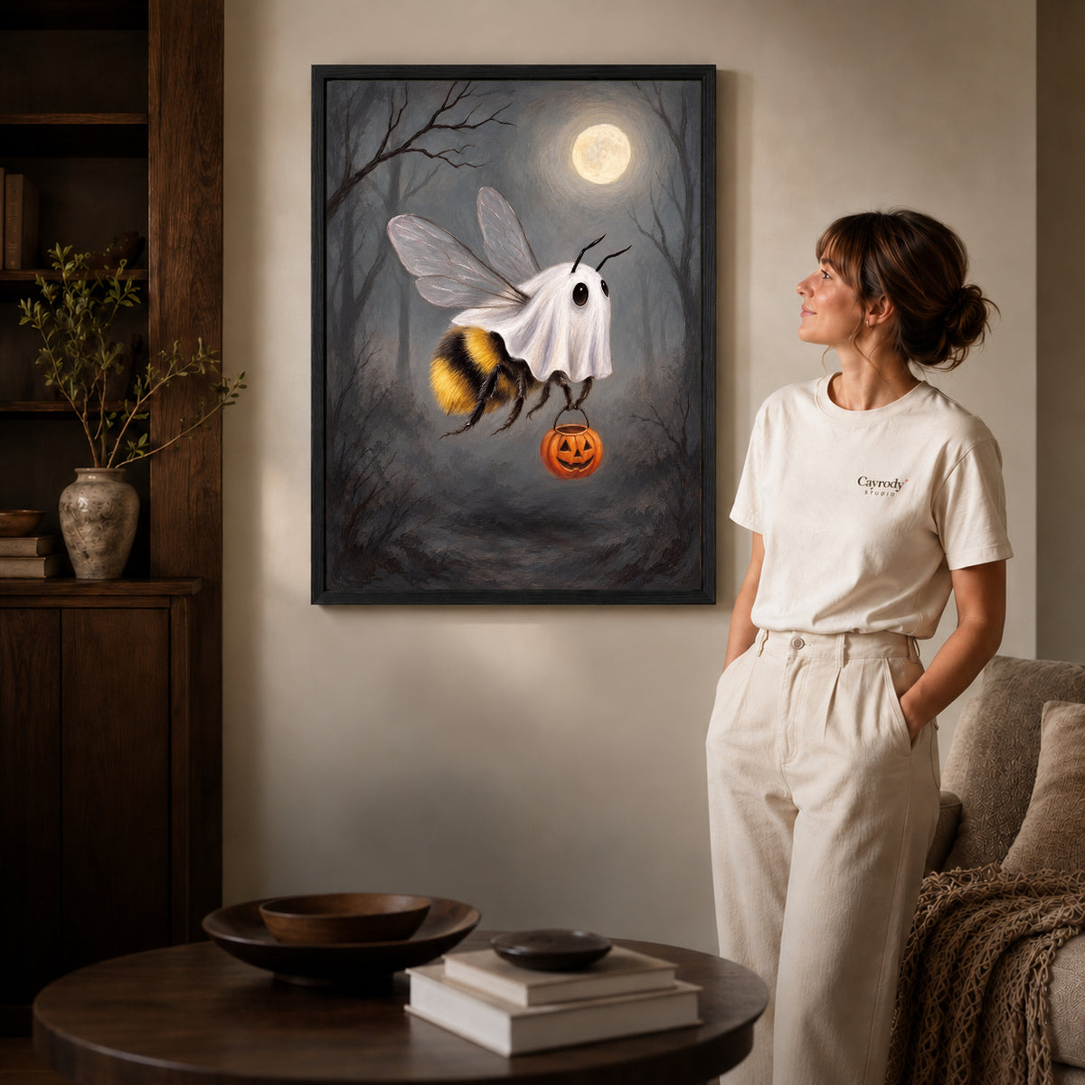 Black framed ghost costume bee canvas art displayed beside a woman in a warm neutral living room.