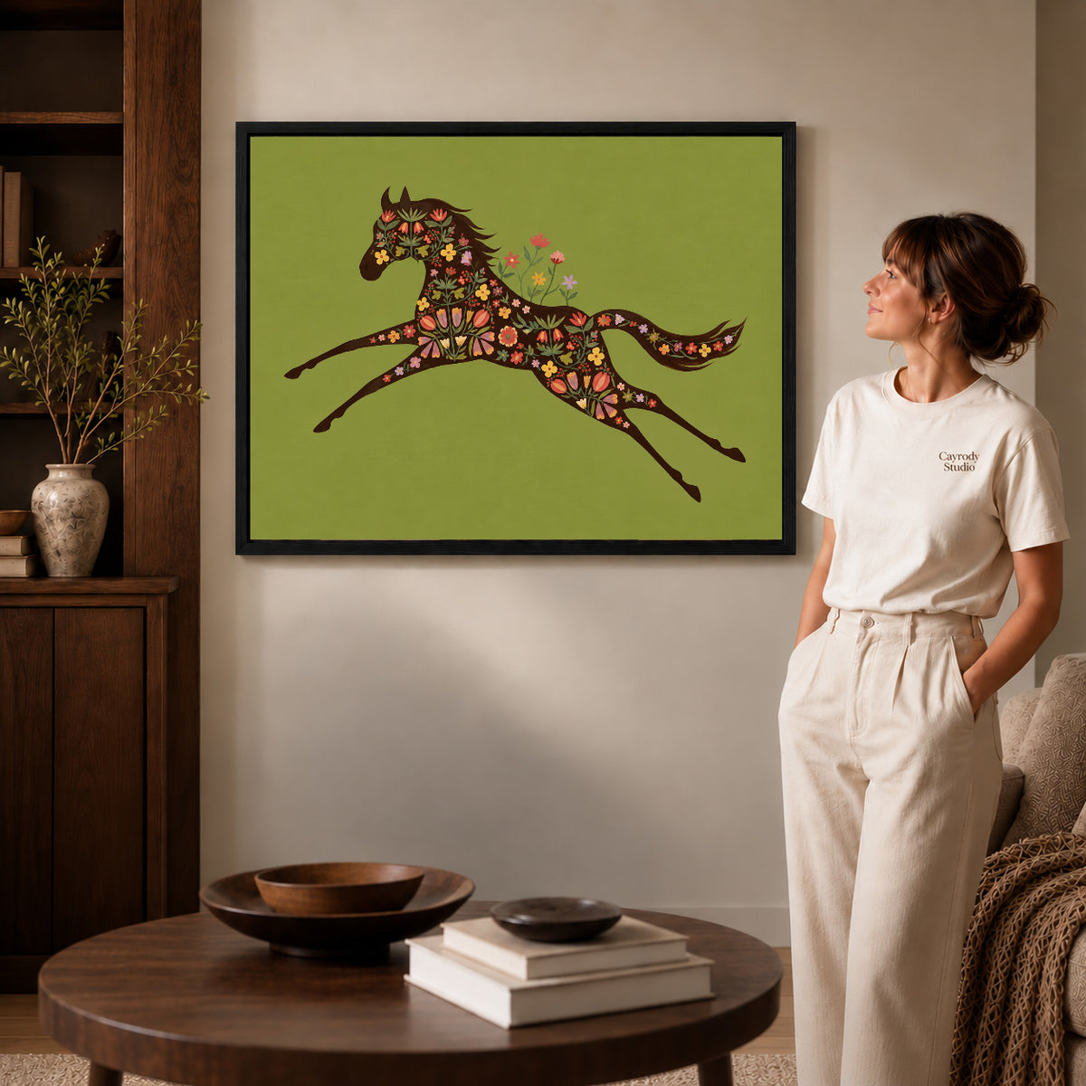 Black framed floral galloping horse artwork beside a woman in a warm neutral living room.