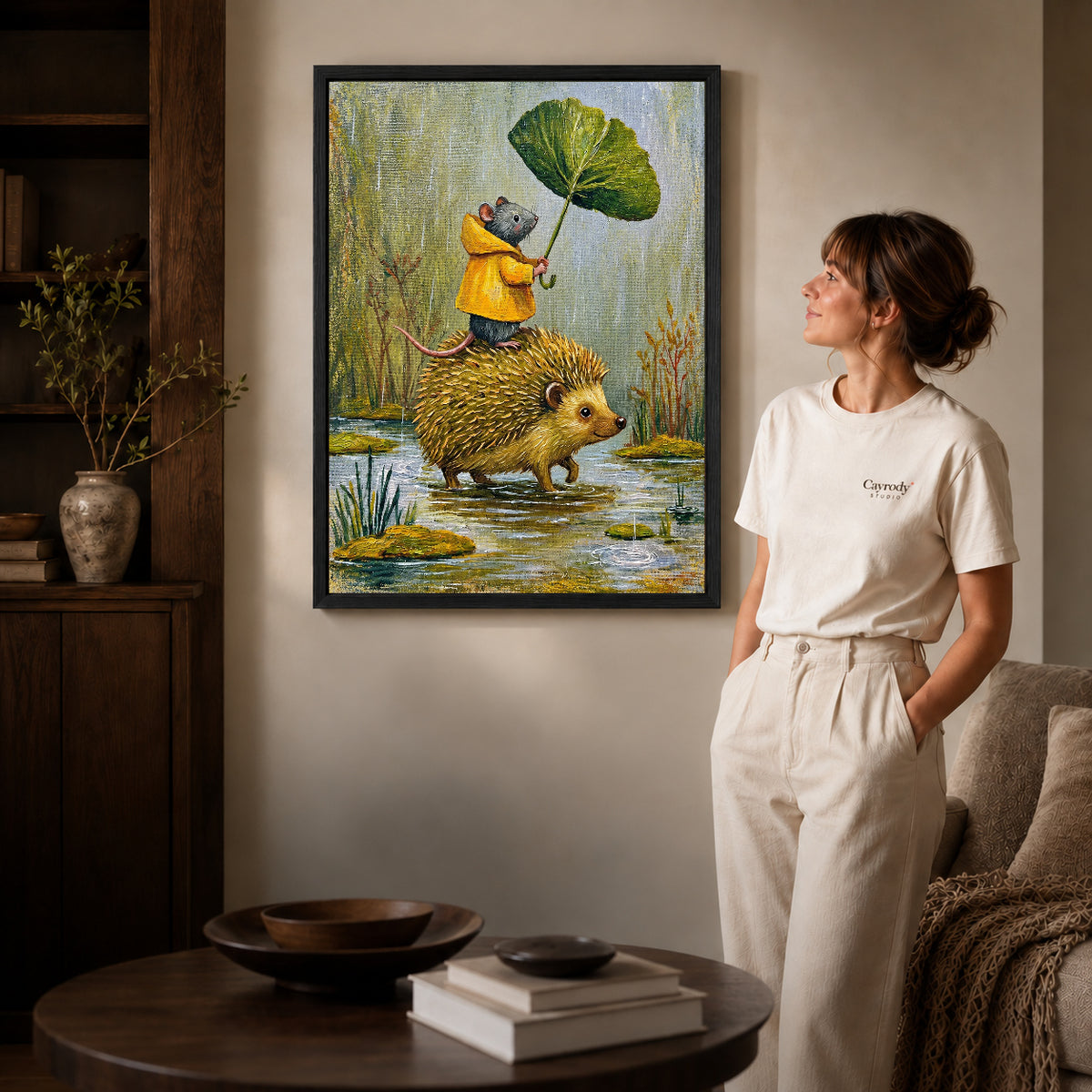 Black framed raincoat mouse ride artwork beside a woman in a warm neutral living room.