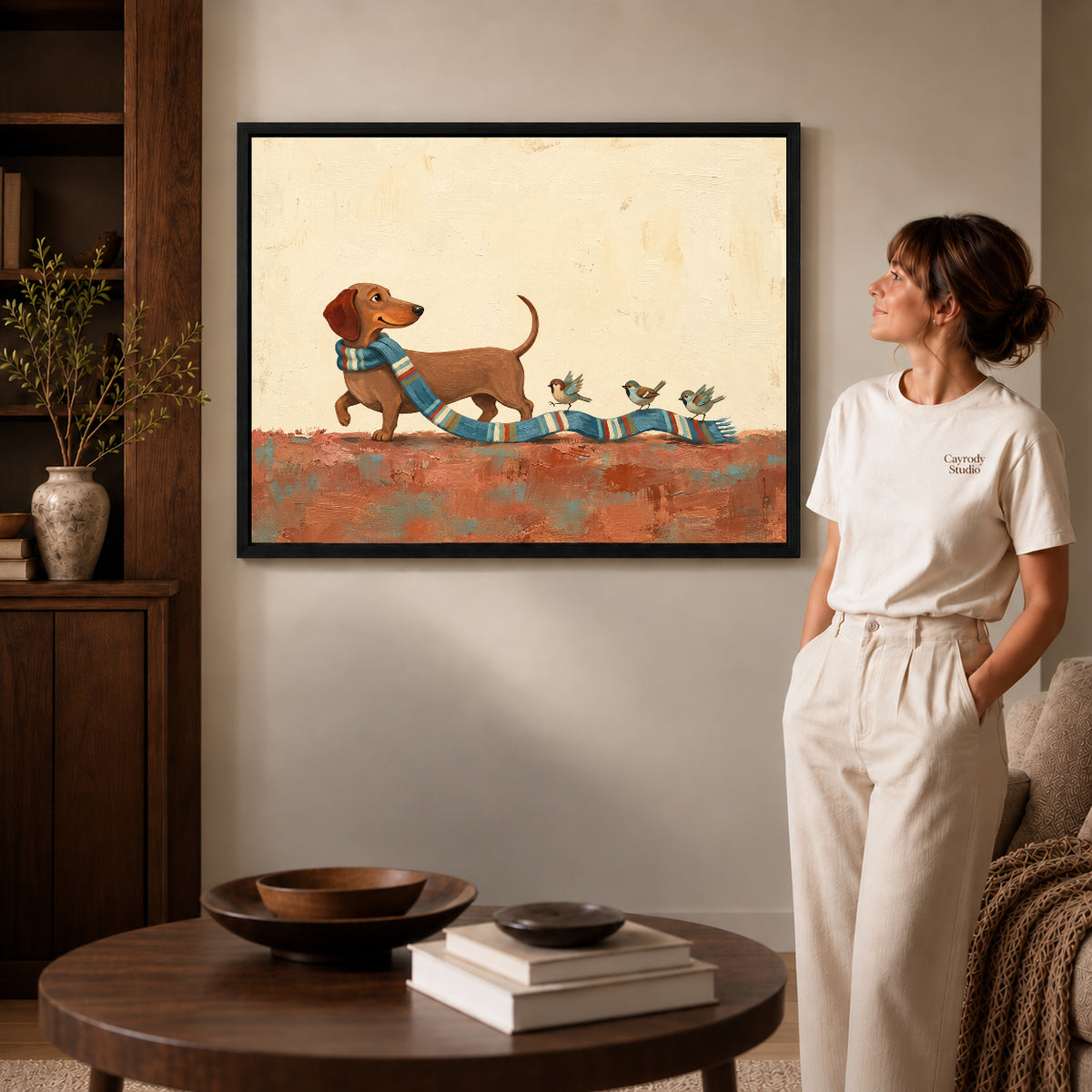 Black framed scarf bird parade artwork beside a woman in a warm neutral living room.