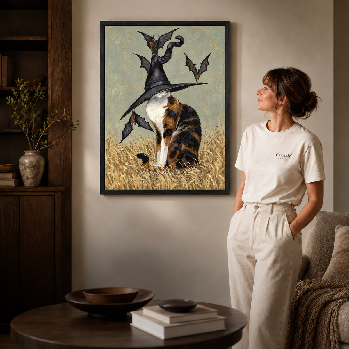 Black framed calico witch hat artwork beside a woman in a warm neutral living room.