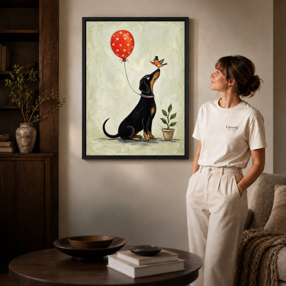 Black framed balloon bird dachshund artwork beside a woman in a warm neutral living room.