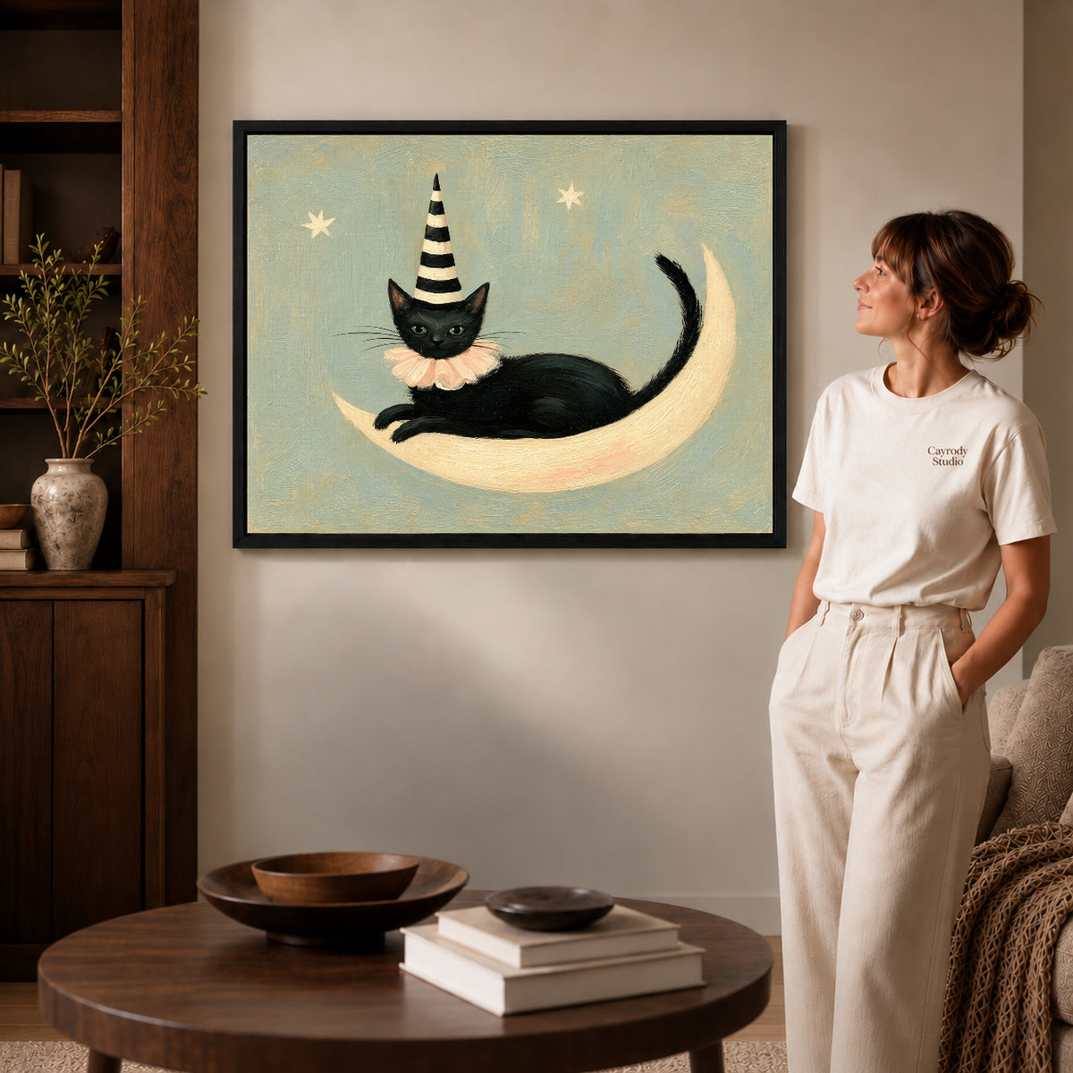 Black framed crescent lounging cat artwork beside a woman in a warm neutral living room.
