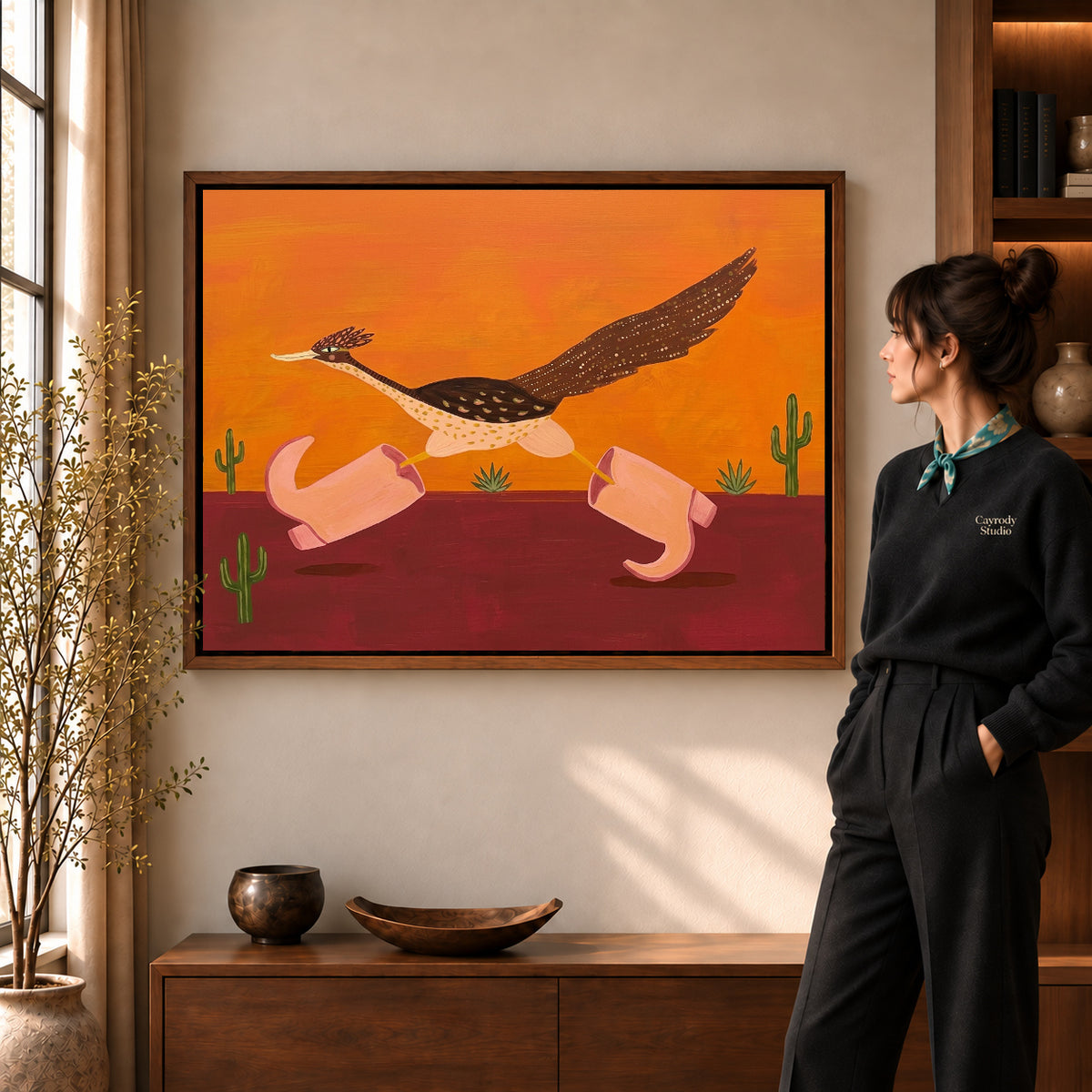 Folk roadrunner pink boots canvas art in a walnut frame beside a standing woman in a warm living room.
