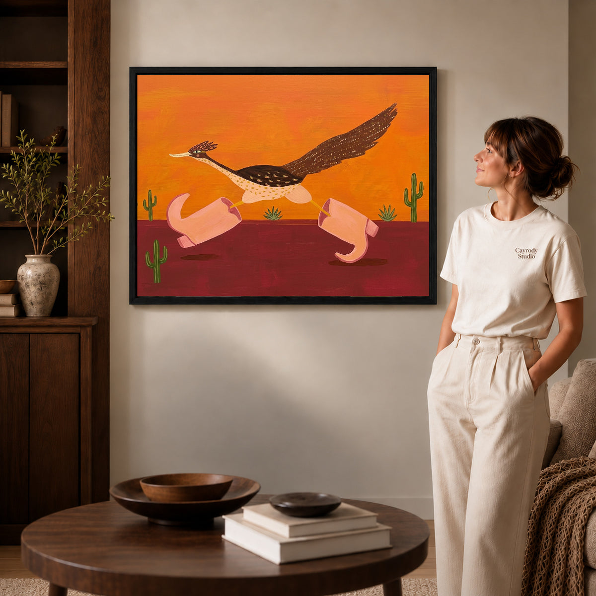 Black framed folk roadrunner pink boots canvas art displayed beside a woman in a warm neutral living room.