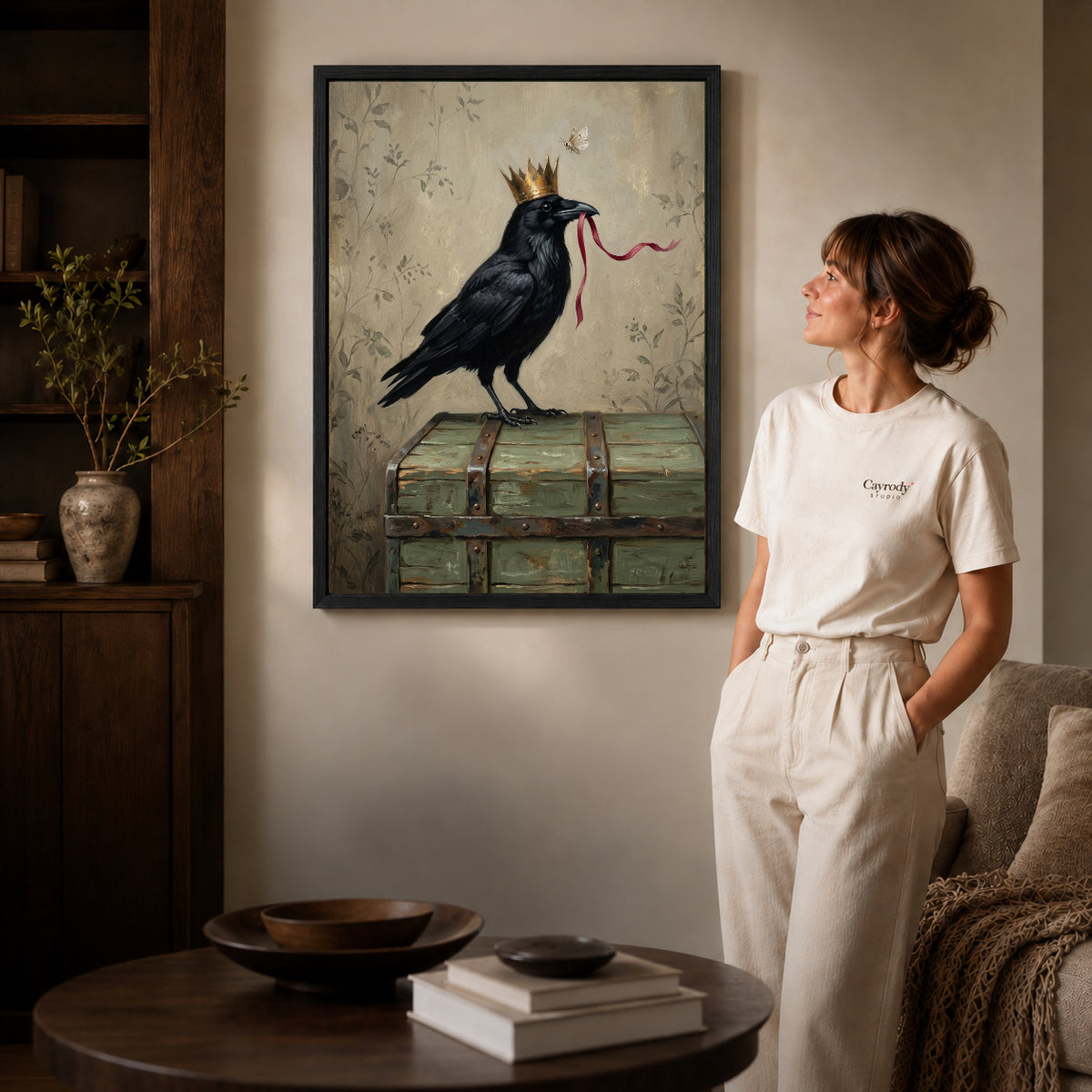 Black framed crowned raven ribbon canvas art displayed beside a woman in a warm neutral living room.