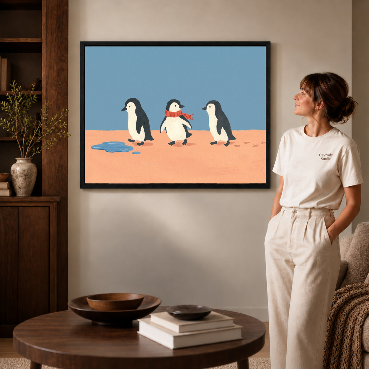 Black framed three penguin friends canvas art displayed beside a woman in a warm neutral living room.