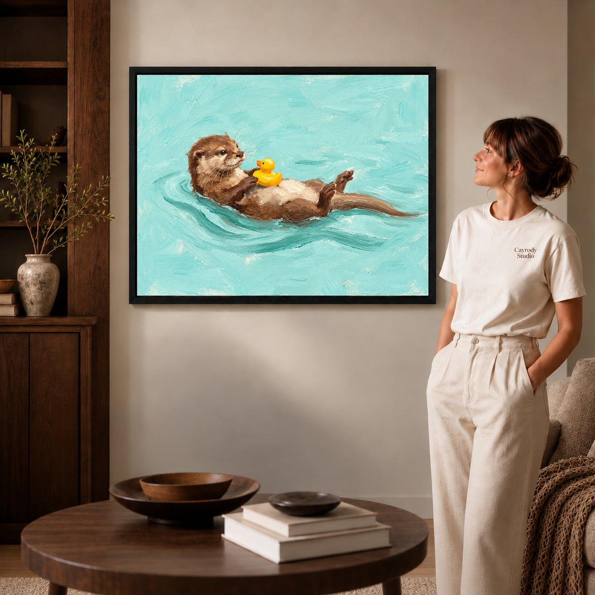 Black framed floating otter duck artwork beside a woman in a warm neutral living room.