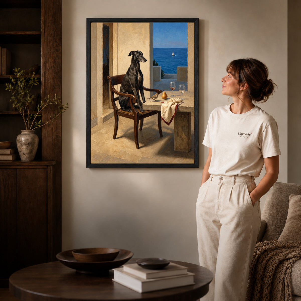 Black framed seaside greyhound portrait artwork beside a woman in a warm neutral living room.