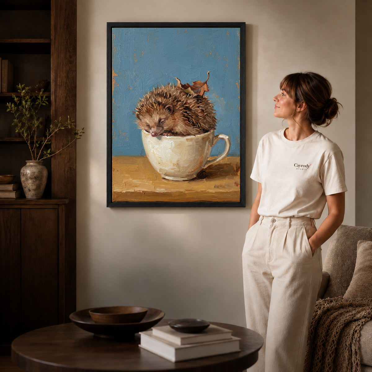 Black framed sleepy teacup hedgehog artwork beside a woman in a warm neutral living room.