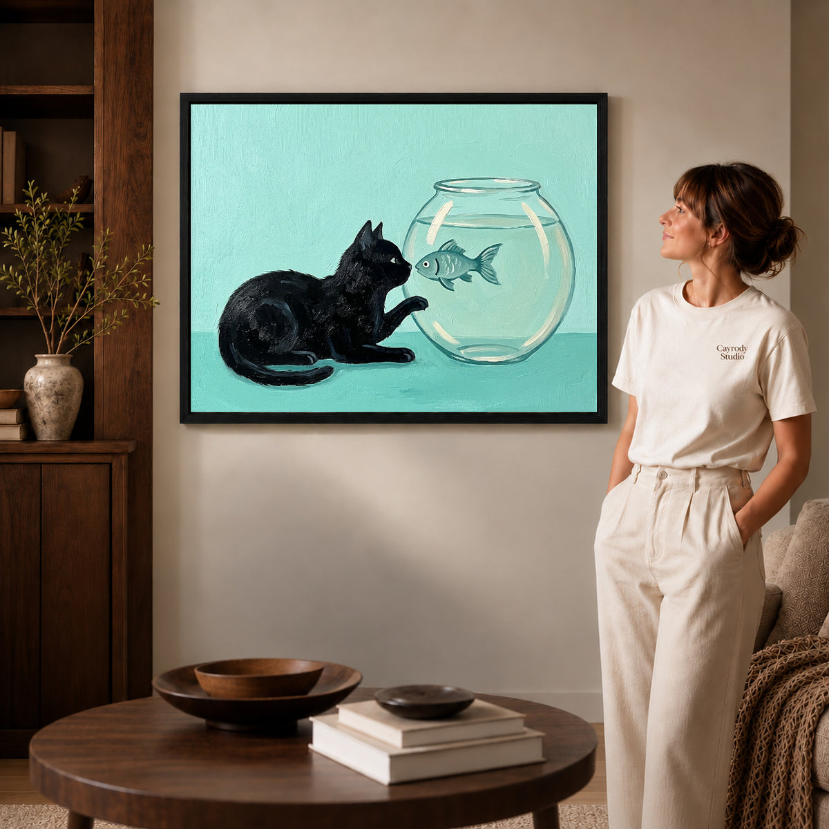 Black framed curious fishbowl cat artwork beside a woman in a warm neutral living room.