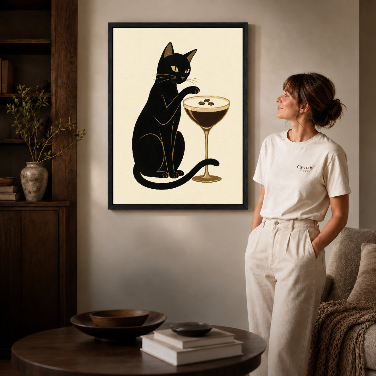 Black framed espresso martini cat artwork beside a woman in a warm neutral living room.
