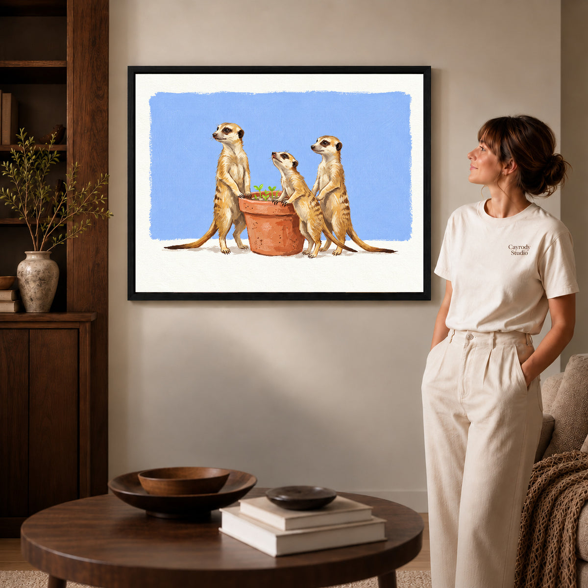 Black framed meerkat gardeners artwork beside a woman in a warm neutral living room.