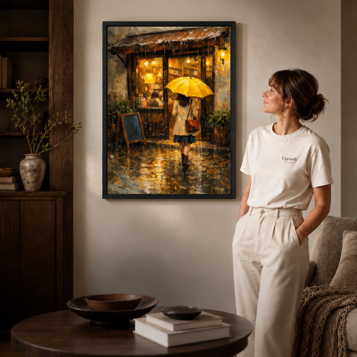 Black framed rainy cafe walk artwork beside a woman in a warm neutral living room.