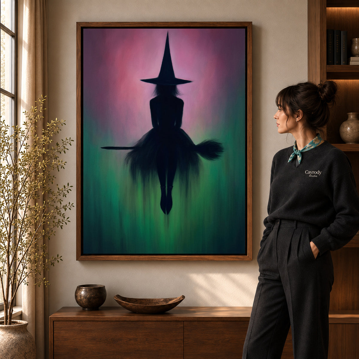 Moody Witch Silhouette artwork in a walnut frame beside a standing woman in a warm room.